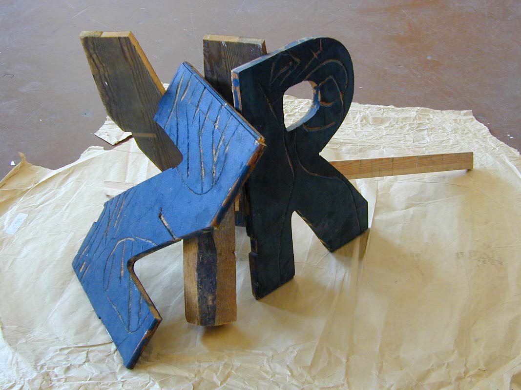 Three wooden letter types covered with black paint standing on the floor 
and leaning against each other, V, R, Z.